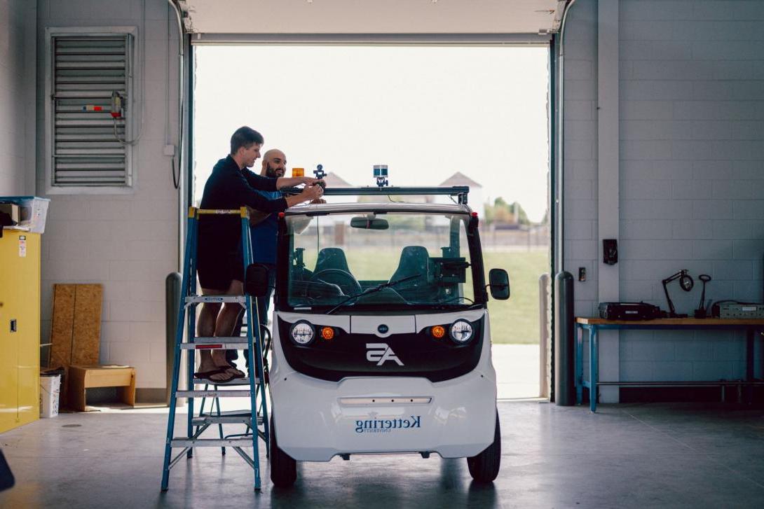 Two Kettering students standing on ladders adjust cameras on top of an electric vehicle in the Mobility Research Center.