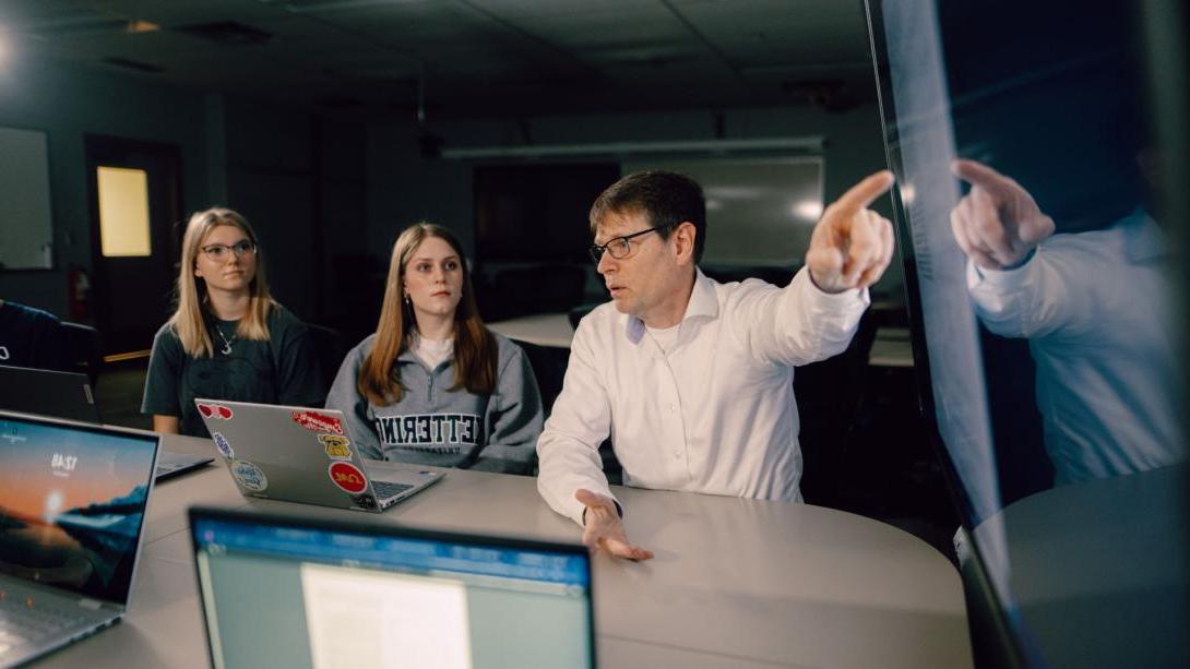 A Kettering professor points at a large television screen while two students look on. Each student has a laptop